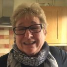 Paula smiling. She's wearing a dark top and grey and white scarf. She has short blond hair and glasses.